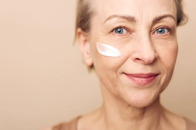 Mature woman with blue eyes applies white cream on cheek, smiling gently against beige background. Concept of skincare, aging care, natural beauty, healthy skin, and self-care routine.
