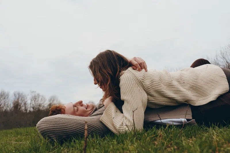 A tender outdoor moment between two adults on a grassy field, one resting atop the other in a warm knit sweater. Natural light, candid and cozy, capturing support, companionship, and calm in a simple