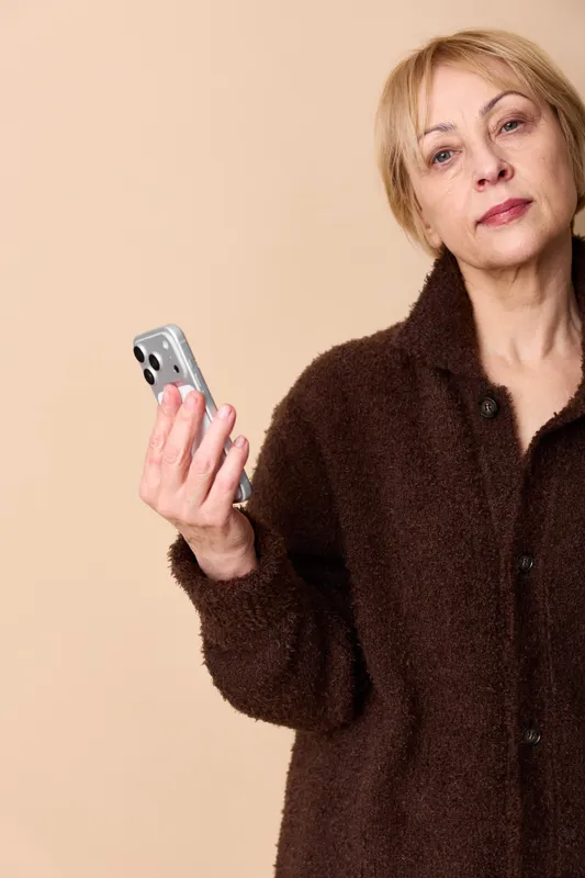 Woman elderly holding smartphone, technology portrait indoors, focused expression, beige studio background, dark coat, mobile communication, calm mood, soft light