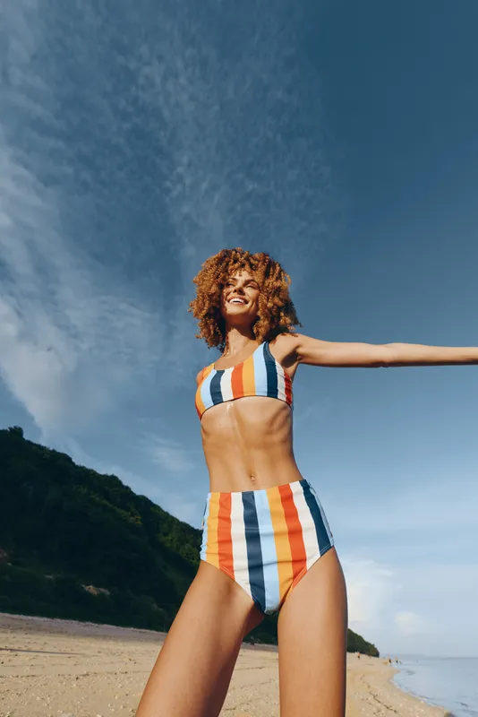 Young woman in striped swimsuit stands on a sandy beach, smiling under clear blue sky. Outdoor summer vacation mood, sunlight on skin, confident pose, calm ocean background.