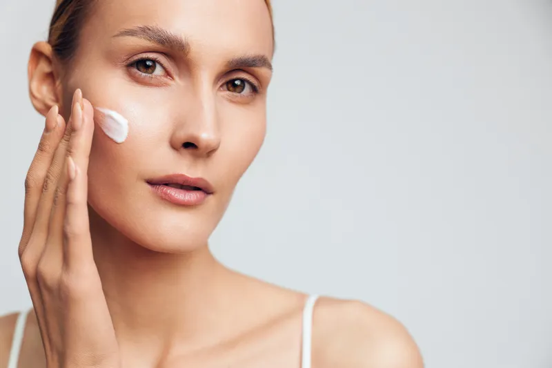 Young woman with clear skin applying white face cream on cheek. Close-up portrait on light background. Concept of skincare, beauty, and natural wellness. Soft lighting highlights healthy skin.
