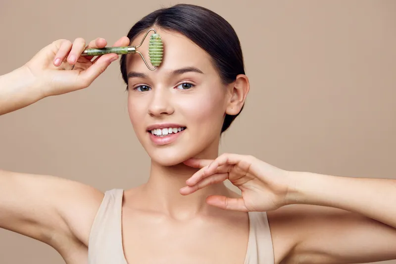Young female model using a jade roller for skincare, showcasing healthy skin and a joyful expression against a neutral background, emphasizing beauty and wellness