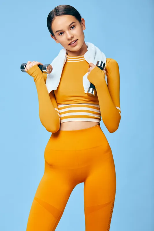 Fitness oriented woman in bright orange activewear poses with a dumbbell and towel, set against a blue backdrop. Dynamic, strong, and stylish athletic scene emphasizing effort, posture, and