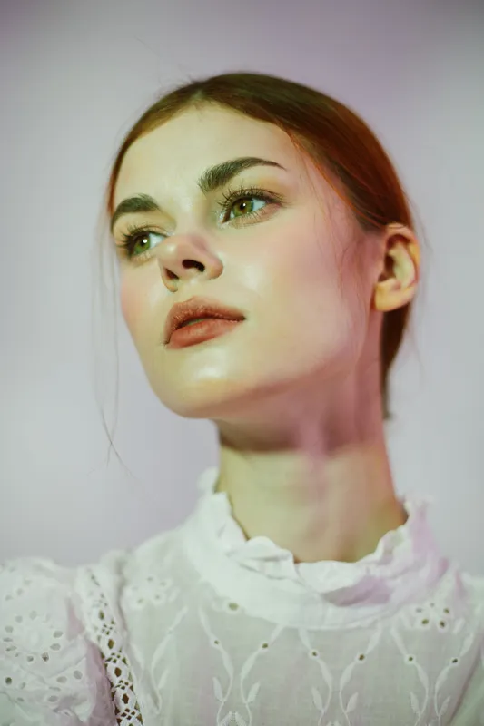 A young woman with long hair, wearing a white lace blouse, gazes thoughtfully against a soft pastel background, conveying elegance and serenity