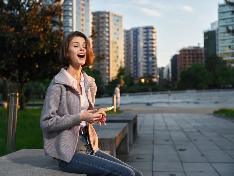 A cheerful young woman sits on a concrete bench in an urban park, texting with a smile, enjoying a pleasant moment outdoors under bright afternoon light, with a modern cityscape behind.