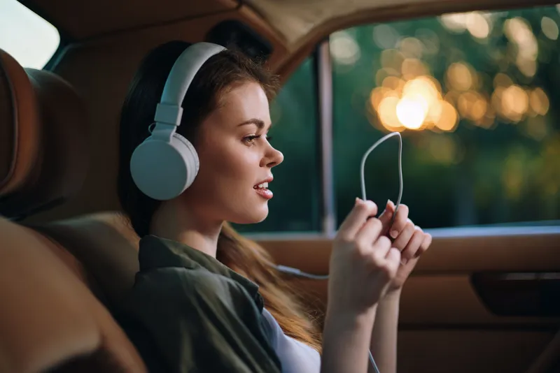 Young woman enjoying music with headphones in a car, relaxed and smiling, surrounded by a warm sunset background, conveying happiness and tranquility.