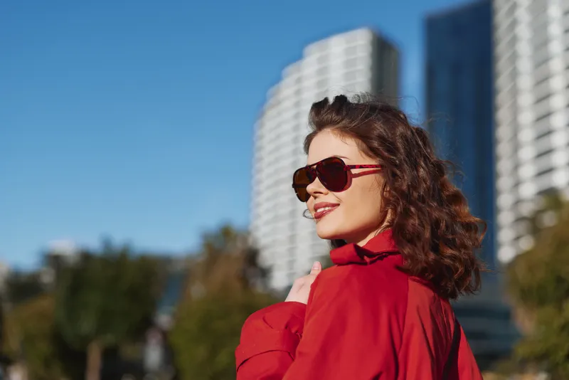A confident woman in a red coat and sunglasses smiles outdoors with urban buildings in the background on a sunny day. Concept for fashion, lifestyle, and city living.
