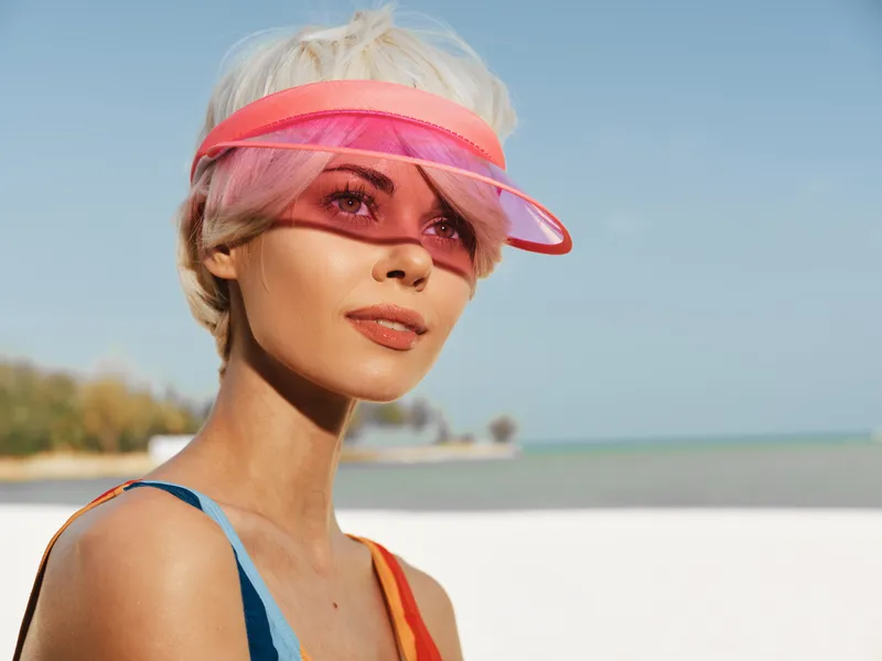 Portrait of a stylish woman on a sunny beach, wearing a pink visor and colorful straps, with natural makeup, highlighting modern summer fashion and outdoor glamour.