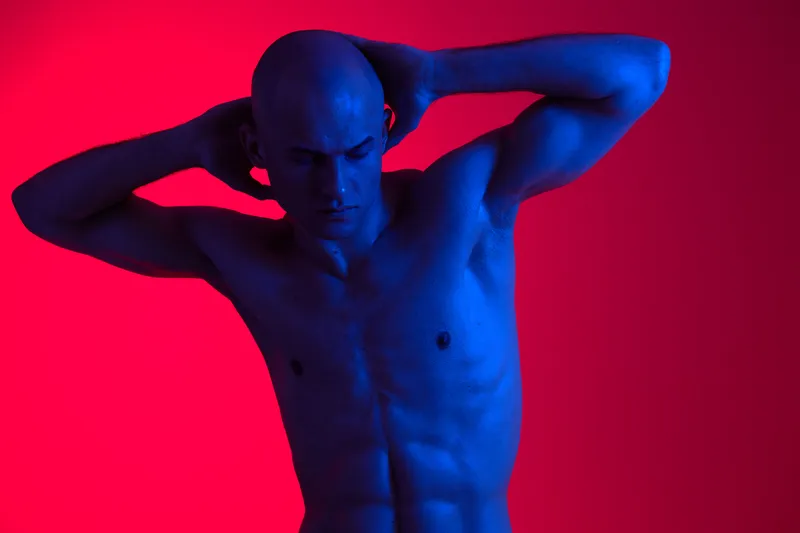 A striking blue-toned image of a man posing with his hands behind his head, showcasing strong muscles and an intense expression against a vibrant red backdrop.