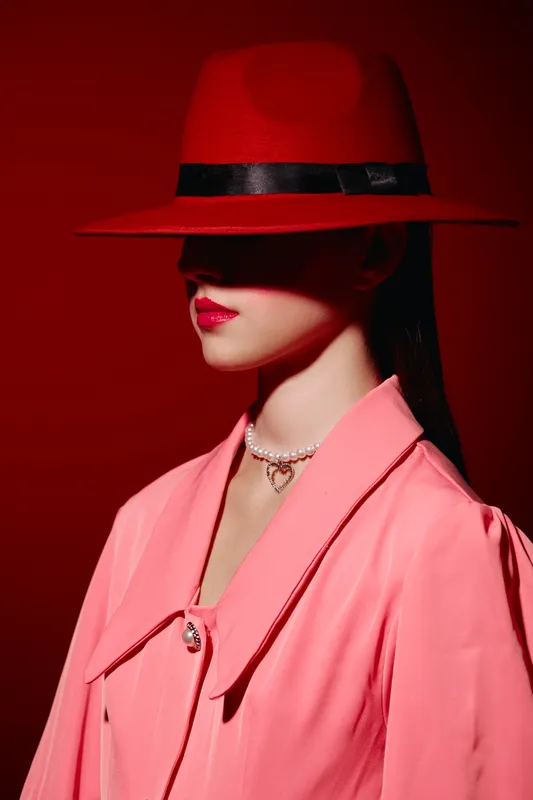 Woman in a stylish red fedora hat and pink coat stands against a red background. Her face is partly covered, creating a mysterious and fashionable beauty portrait concept.