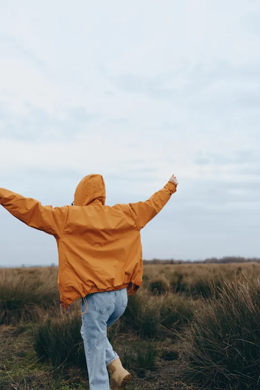 Woman outdoors lifestyle in grassy field, orange jacket hood, blue jeans, arms open wide, daylight sky, relaxed stance, wind moving fabric, natural colors, freedom moment, candid portrait