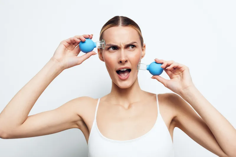 Young woman with clear skin holds blue facial suction cups near her face, expressing surprise. White background, skincare concept with beauty and self-care focus.