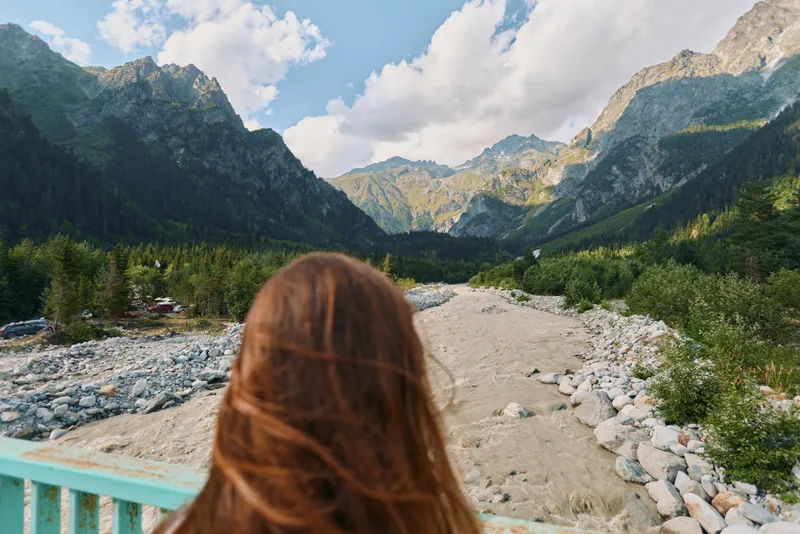 A person with long brown hair stands on a bridge overlooking a rocky river valley framed by towering mountains. They gaze outward, enjoying tranquil natural scenery and peaceful solitude.