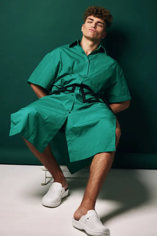Young man sitting on chair wearing green oversized shirt dress with black belt and white slip on shoes against dark green background in studio setting.