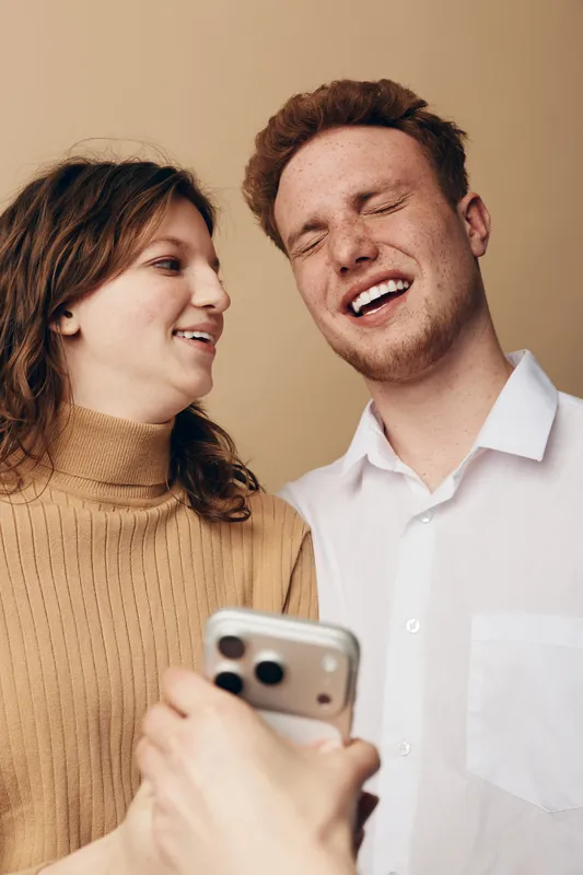Young man and woman smiling and laughing while looking at a smartphone. Casual indoor setting with warm lighting and beige background. Concept of friendship and happiness.