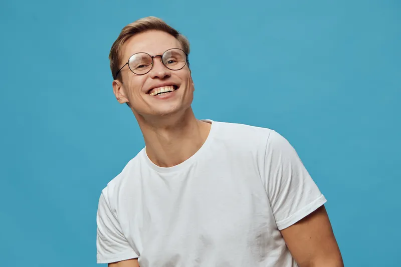 Smiling young man wearing glasses and casual white t-shirt looking happy against a plain blue background, studio portrait, positive expression, cheerful mood, lifestyle concept, human emotion, joyful 