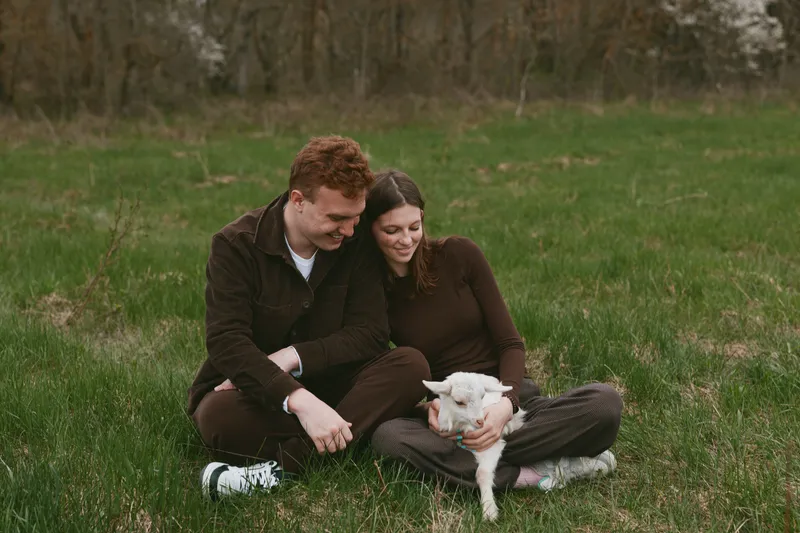 A couple sits in a field with a curious goat kid, enjoying outdoors and grass as a gentle lifestyle moment highlighting bonding and shared joy in a warm, earthy scene that captures connection, care,