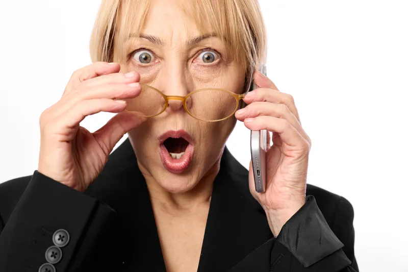 Shocked woman in business suit, wide eyes and surprised expression, adjusting glasses with fingers, holding smartphone phone to ear, hands near face, white studio background