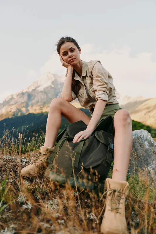 Young woman sits on a rocky mountainside with a backpack and boots, wearing khaki shirt, as sunlit meadow and distant peaks frame her thoughtful, relaxed moment