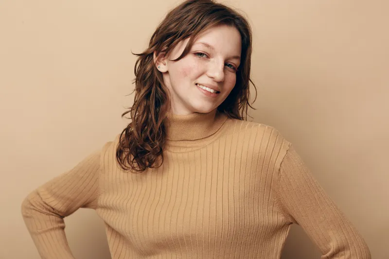 Smiling young woman with wet hair wearing a beige knitted sweater poses confidently against a matching beige background. Concept of casual style, natural beauty, and autumn fashion.
