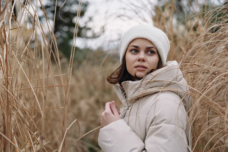 Winter scene featuring a woman in a beige coat and white knit hat among tall grasses, natural backdrop, calm mood, thoughtful gaze, outdoor solitude and cool air.