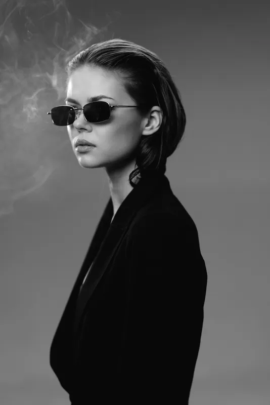 Black and white portrait of a stylish woman wearing sunglasses and a dark blazer with a serious expression and smoke in the background