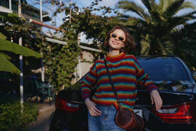 Woman in her late 20s wearing a colorful striped sweater and yellow sunglasses, smiling while leaning against a black car with a brown crossbody bag in tropical outdoor street.