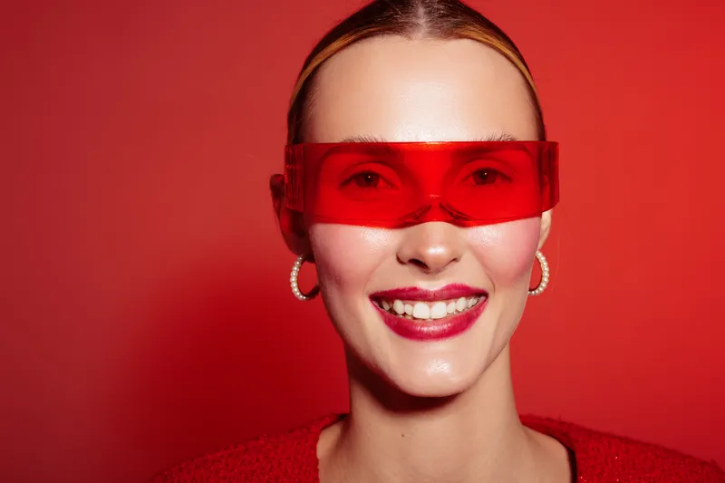 Smiling woman with red transparent glasses and hoop earrings poses on red background. Fashion portrait with bright makeup and slicked back hair. Concept of style and modern accessories.