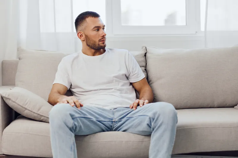 Thoughtful man sitting on a cozy sofa, reflecting in a serene indoor environment with natural light. Stylish outfit complements modern decor and calm atmosphere.
