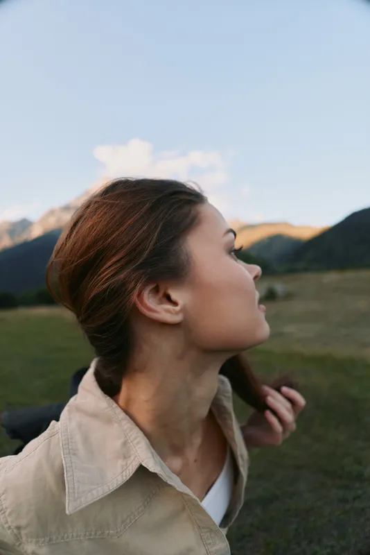 Young woman with brown hair enjoys outdoor lifestyle in nature, looking sideways with peaceful expression, wearing casual jacket in mountain landscape.