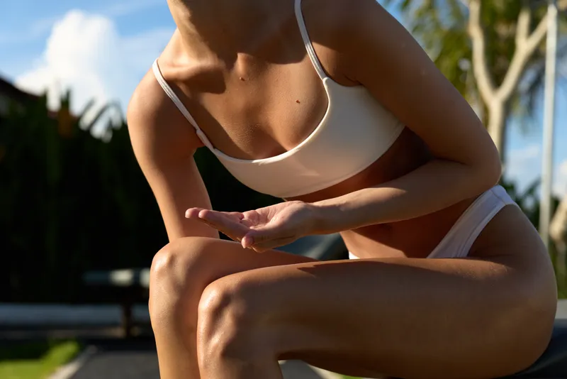 A sunlit outdoor scene shows a fit woman in a white bikini crouching beside a pool, with lush greenery and blue water in the background. swimwear outdoor summer fitness pose toned