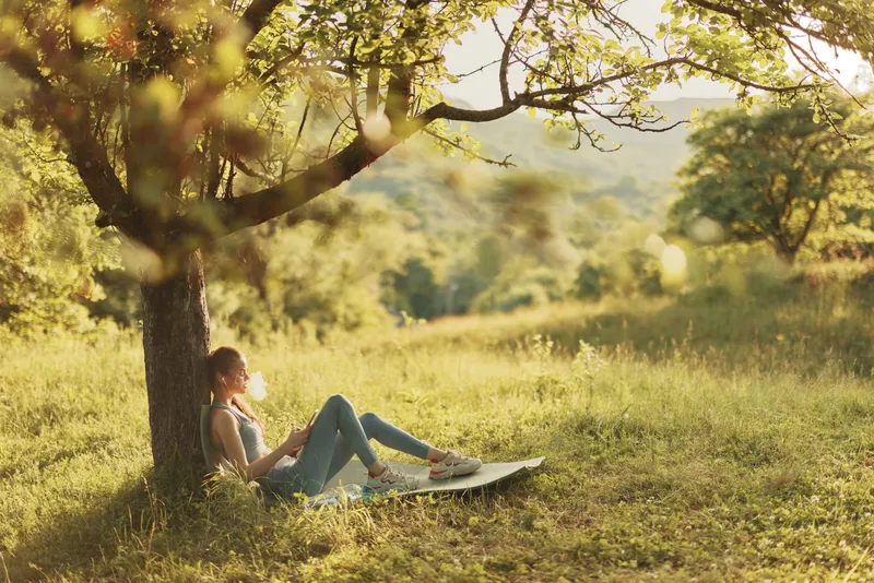 Girl relaxing under a tree in a green meadow, enjoying a book on a sunny day, representing tranquil moments in nature and healthy leisure