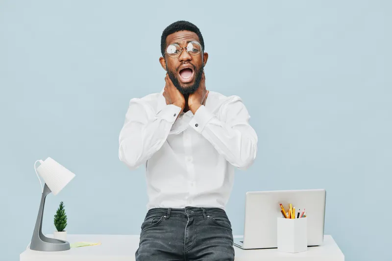Frustrated young black man in white shirt holding his face with both hands, expressing shock or disbelief, sitting at modern desk with laptop, pencil holder, desk lamp, and small plant against plain b