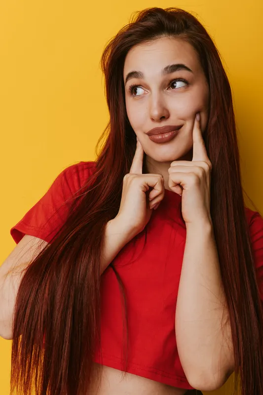 Young woman with long hair and red shirt smiling while touching her cheeks with fingers, looking sideways on yellow background. Portrait, emotion and casual style with natural light.