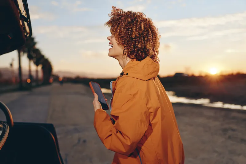 Woman with curly hair portrait in orange coat walking outdoors at sunset, holding smartphone in hands, looking to side with smile, lifestyle scene, warm golden light on quiet street