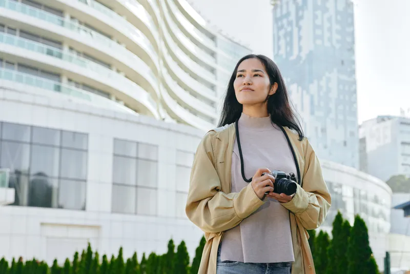 A woman stands outdoors with a camera under a modern cityscape, wearing a beige coat and soft sweater, gazing ahead as she captures scenes of urban life and architecture with style and momentum.
