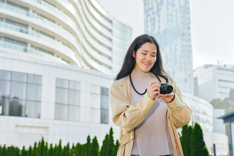Camera in hand, smiling woman outdoors in an urban setting with modern buildings behind, casual jacket, bright daylight, candid lifestyle shot suitable for travel, fashion or media projects