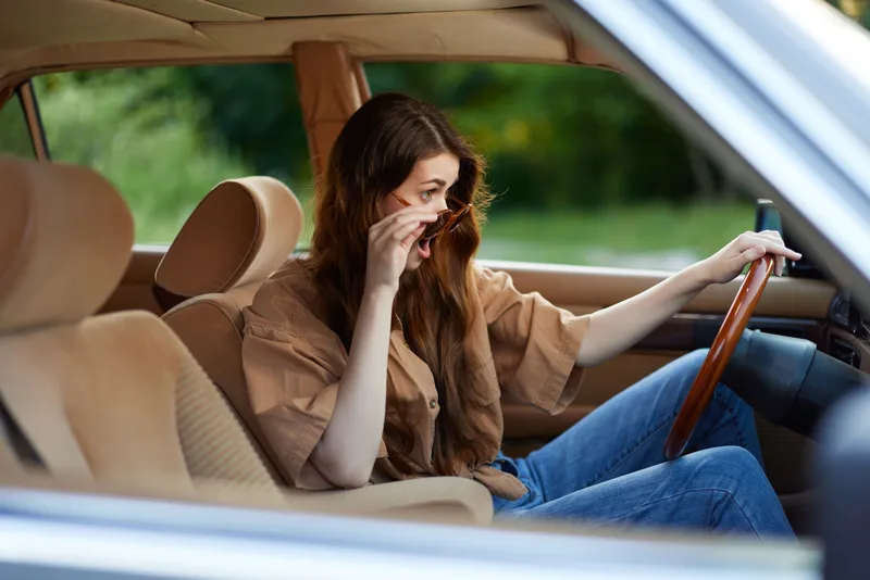Young woman with long hair, wearing a casual brown shirt, expresses concern while sitting in a car, surrounded by a lush green background.