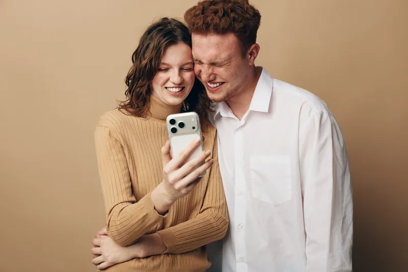 Smiling young couple takes selfie with smartphone, standing close together on beige background. Concept of friendship, technology, happiness, and casual lifestyle in studio setting.