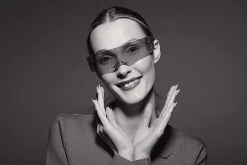 Smiling white woman with pale skin wearing sunglasses and a sweater poses with hands near face on dark background.