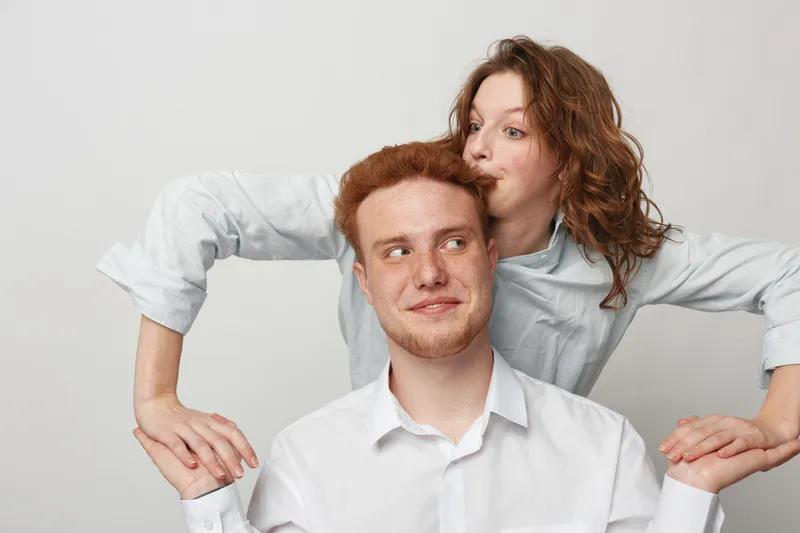 Young redhead man and woman with curly hair share a playful moment, holding hands and smiling against a plain light background. Concept of friendship and happiness.
