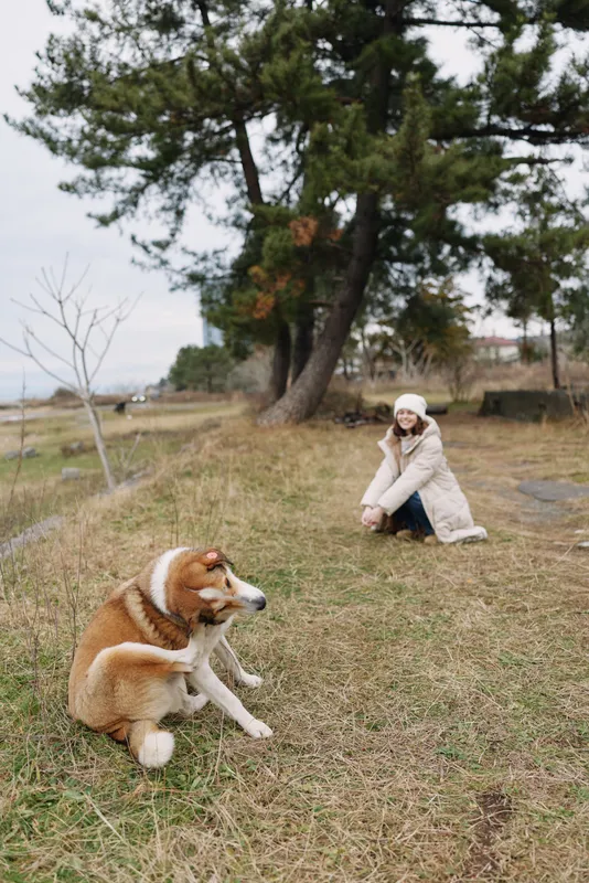 Woman in a light coat kneels in a park with a friendly dog, outdoor nature and trees, capturing a calm moment in a natural setting.