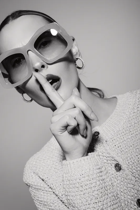 Stylish caucasian woman with light skin and sunglasses making shushing gesture. Close-up portrait in monochrome with knitted sweater and hoop earrings representing secrecy concept.