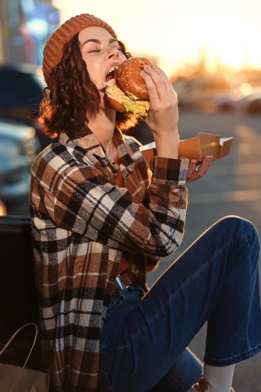 Woman burger eating candid lifestyle authenticity golden urban portrait at sunset, joyful young woman biting a burger outdoors with emotional storytelling vibe and mindful living mood.
