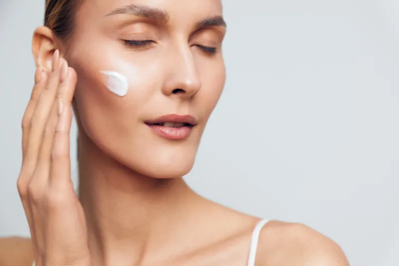 Skincare focused portrait of a woman applying cream to her cheek during a routine, soft studio lighting, calm mood, close up on smooth skin, natural makeup, personal care moment