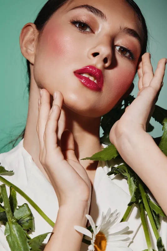Close-up of a fashionable young woman with glossy red lips and glowing skin posing with her hands near her face against a green background. Concept of beauty makeup and fashion creativity.
