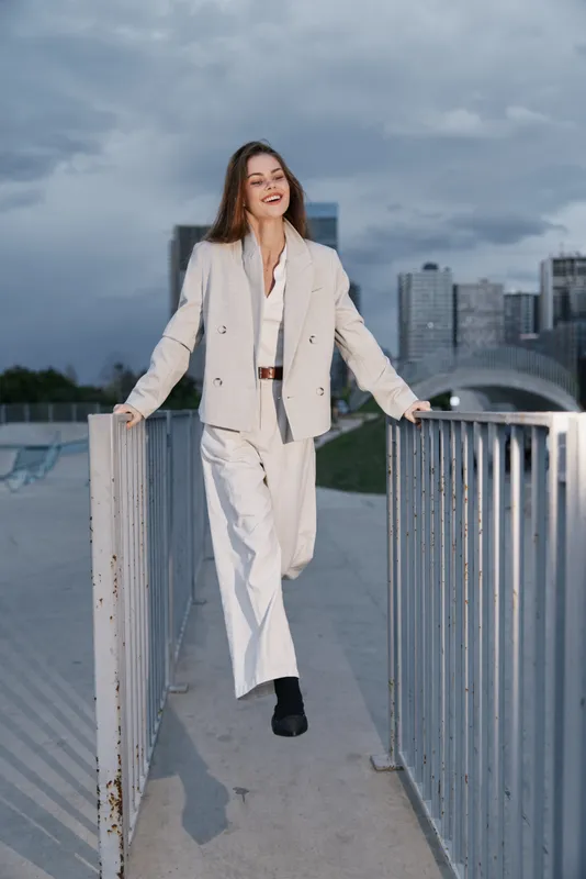 Young woman in beige business suit smiling confidently outdoors with city skyline background, fashion style, urban business concept, modern professional attire