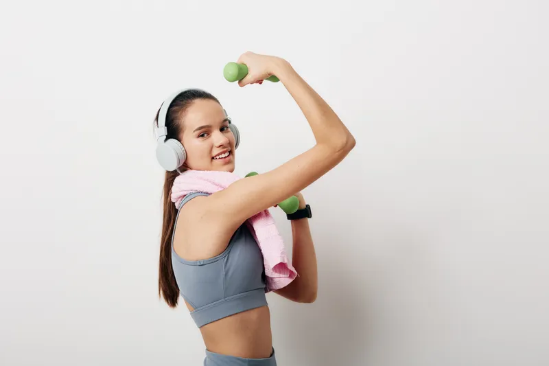 Woman in athletic wear lifts a small dumbbell with headphones on and a towel draped over her shoulders, capturing a bright fitness moment in a clean studio.