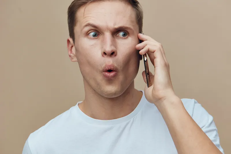 Close-up of surprised young Caucasian man in casual white t-shirt talking on mobile phone with expressive facial expression isolated on plain beige background studio portrait. People lifestyle concept
