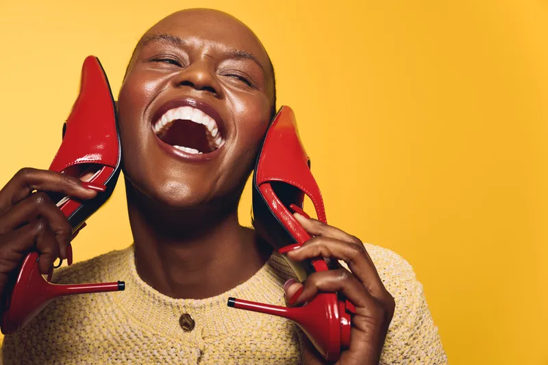 Happy dark skinned African woman laughs holding red high heel shoes near her face. Yellow background, joyful mood, fashion concept with bright colors and female beauty expression.